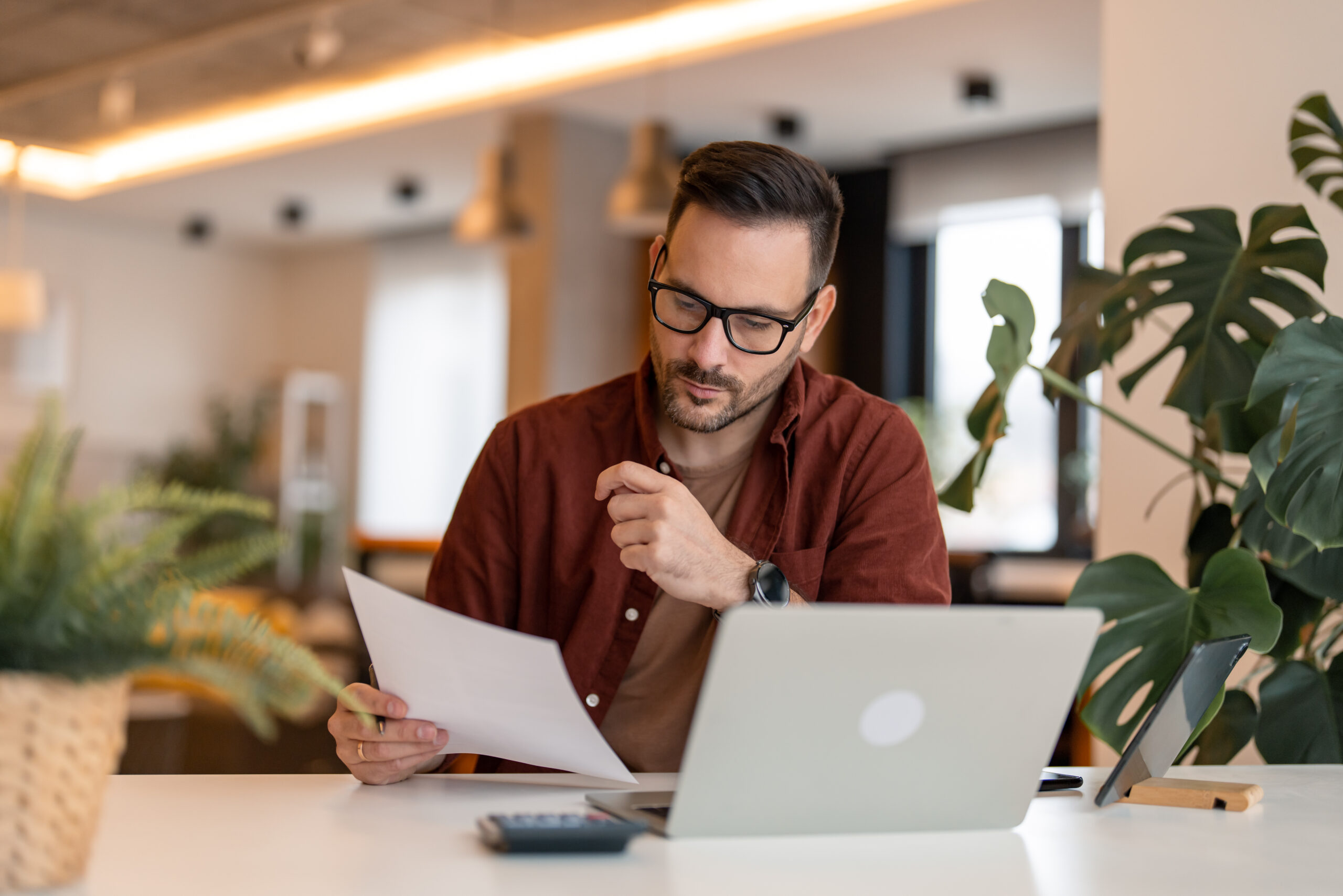 Serious millennial man using laptop sitting at the table in a home office, focused guy in casual clothing looking at the paper, communicating online, writing emails, distantly working or studying on computer at home.