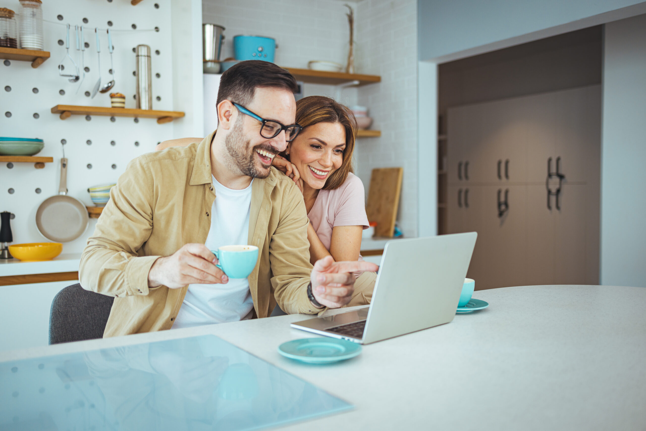 Pleasant family couple sitting at big wooden table in modern kitchen, looking at laptop screen. Happy young mixed race married spouse web surfing, making purchases online or booking flight tickets.