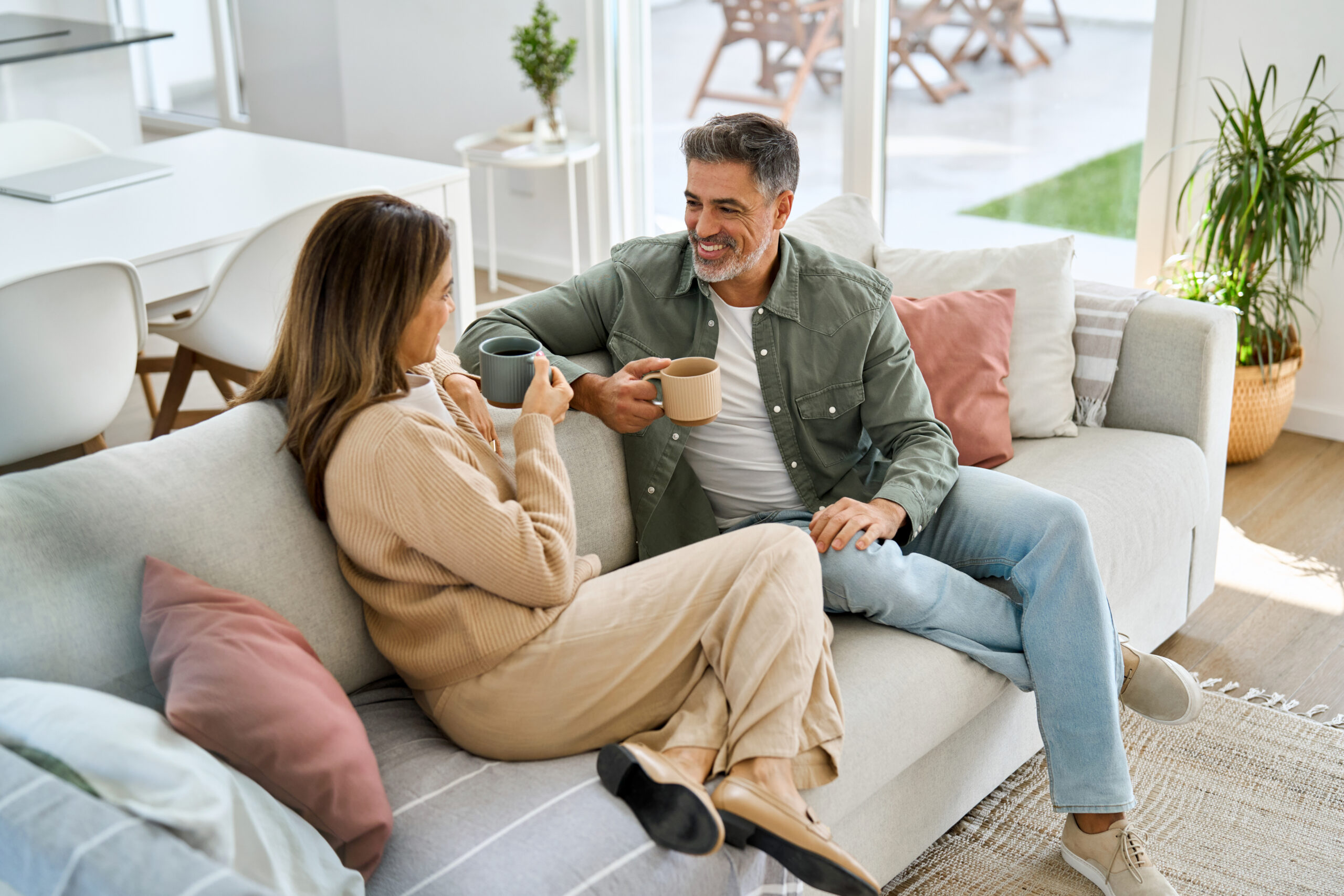 Happy middle aged mature couple talking drinking coffee relaxing on couch at home. Relaxed older man and woman in love sitting on sofa enjoying conversation in living room. Candid photo.