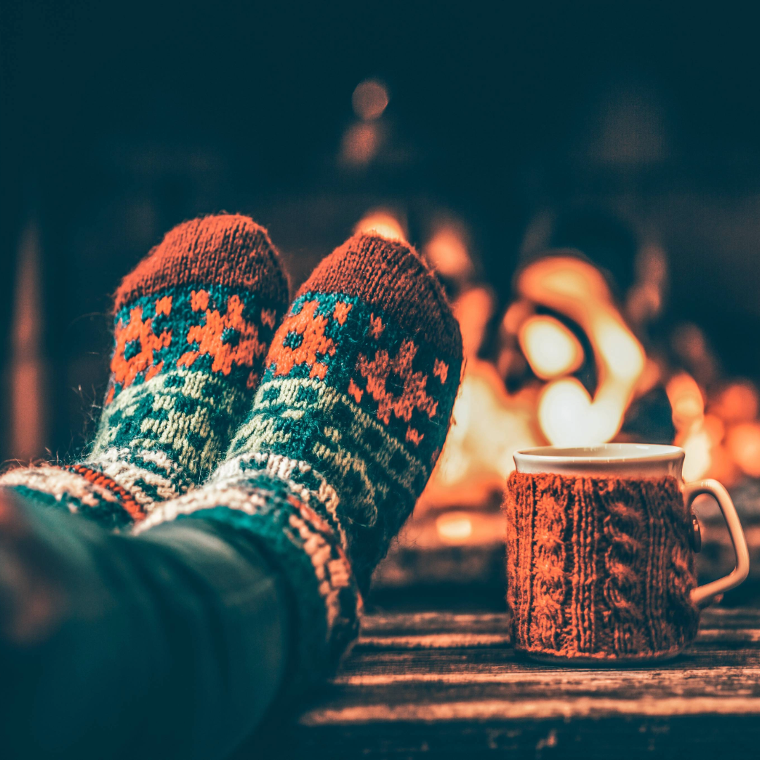 Feet in woollen socks by the Christmas fireplace. Woman relaxes by warm fire with a cup of hot drink and warming up her feet in woollen socks. Close up on feet. Winter and Christmas holidays concept.